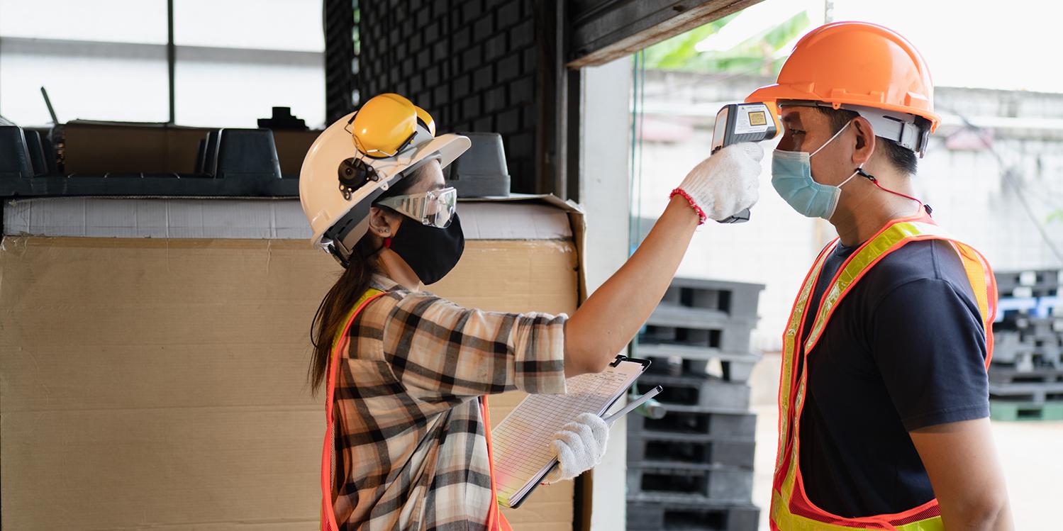 Two factory workers in safety gear and masks, female scanning a male for temperature with a digital thermometer