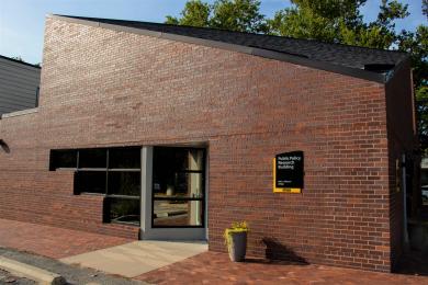 a brick building with a corner office and windows, a plant with yellow flowers sits outside and a sign reads 'Public Policy Research Building' on the wall