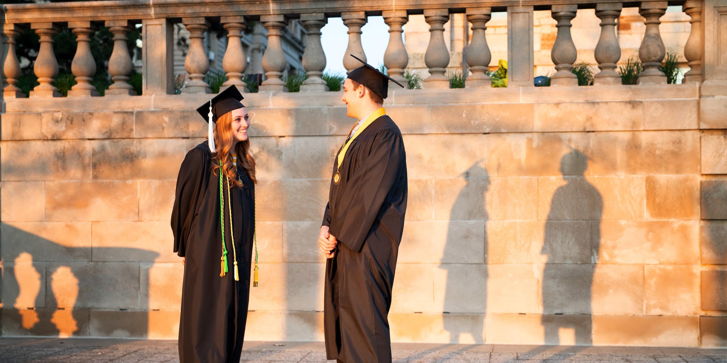 Two students in graduation caps and gowns, standing on the west terrace of the Old Capitol Museum, their shadows cast on the wall behind them.