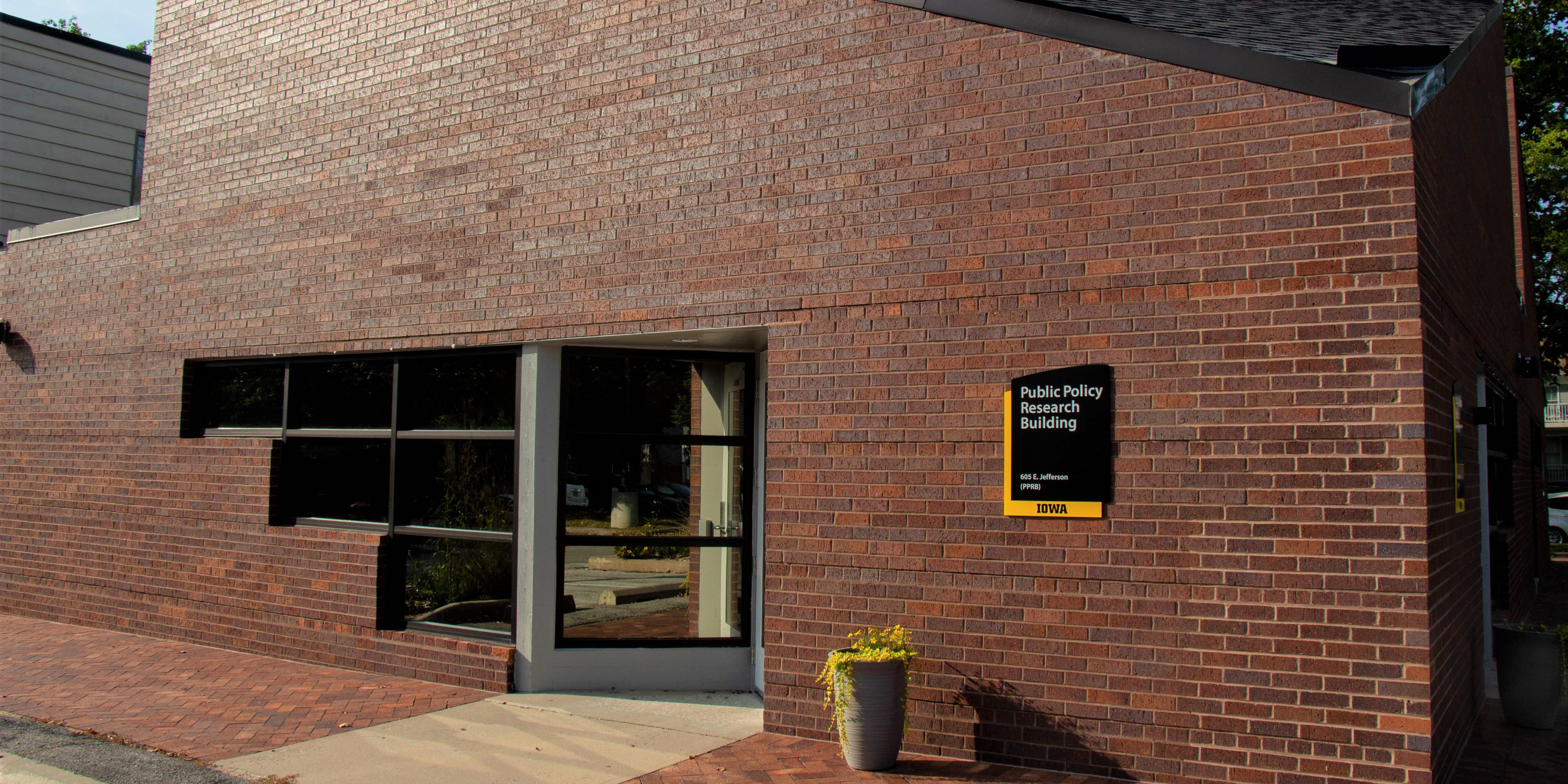 a brick building with a corner office and windows, a plant with yellow flowers sits outside and a sign reads 'Public Policy Research Building' on the wall